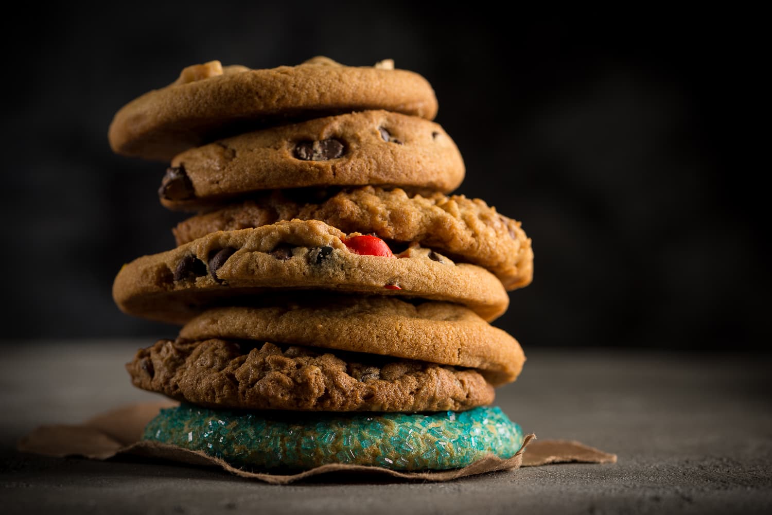 A tall stack of chocolate chip and M&M cookies with a teal sugar cookie at the base