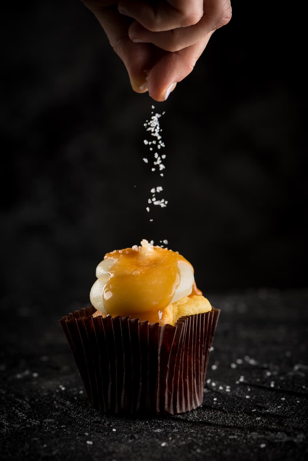 A salted-caramel cupcake with a hand sprinkling sea salt cascading from above, dark and cinematic