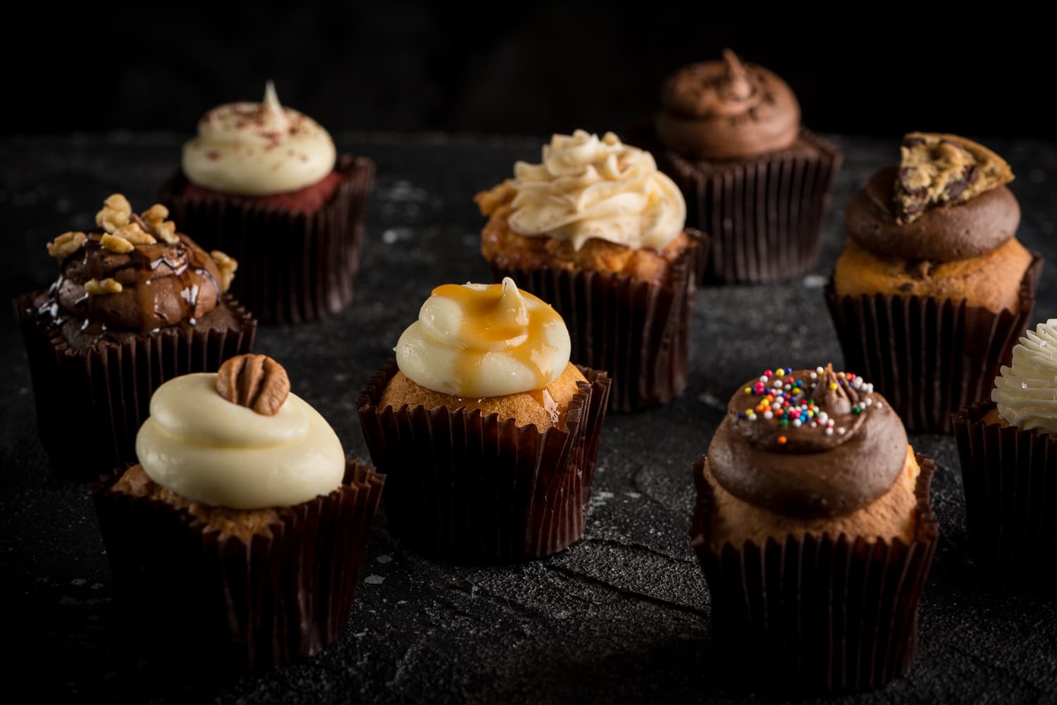 An assortment of Little Cakes With Big Attitude cupcakes on dark stone, Alexandria LA bakery